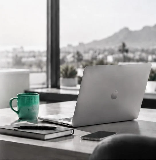 A calm, minimal desk with an open silver laptop, a notebook, a phone and a green ceramic mug, by a window with a soft view of Cape Town