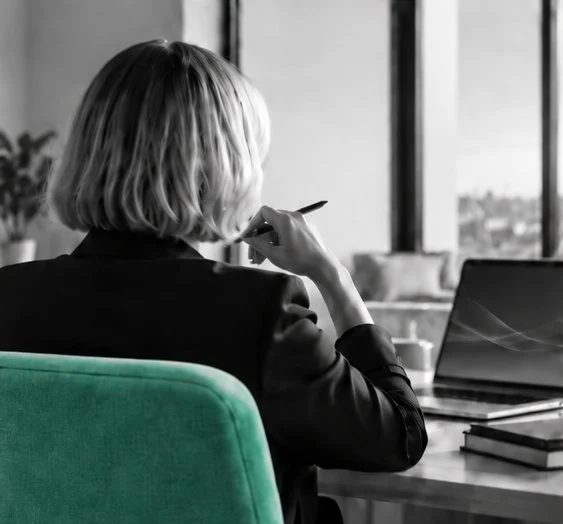 A woman with a blonde bob, seen from behind, thinking at her desk in a green chair